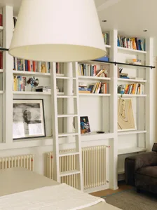 Cozy living room with a white bookshelf, couch, and dining table near a window, featuring a lamp and a comfortable arrangement of books and furniture