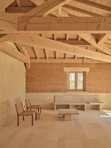 Dining area with a wooden floor, stone wall, dining table, chairs, and bench, lit by natural sunlight through a window