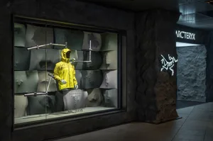 A mannequin wearing a yellow jacket stands in front of a stone wall in a store display, creating a striking contrast