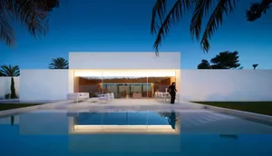 A modern house with a white exterior, surrounded by palm trees, features a pool with chairs and a dining table, creating a luxurious and inviting atmosphere