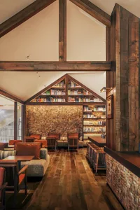 Cozy living room with a high ceiling, wooden elements, and natural light from windows, featuring a large bookshelf and multiple seating areas