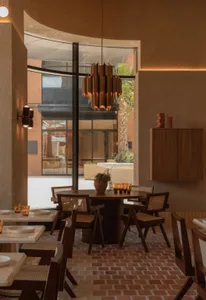 A modern restaurant interior with a dining table, chairs, potted plants, a kitchen with a refrigerator, oven, and sink, and a warm color palette