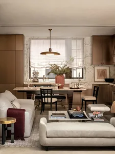 Cozy living room with a couch, chair, and potted plant, adjacent to a dining area with a table and chairs, decorated with books, a vase, and a bowl, featuring a mix of wood and fabric materials and natural lighting