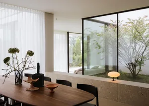 Cozy modern living space with a wooden dining table, chairs, a couch, and a potted plant, lit by natural light from a large window