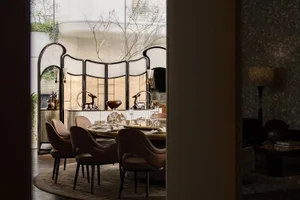 A modern dining room with a large wooden dining table and chairs, set with various items, surrounded by a large window allowing natural light to fill the space