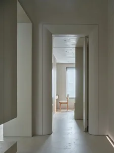 Narrow modern hallway with a white color scheme, concrete walls, and a wooden floor, featuring a chair near the doorway in a dimly lit space