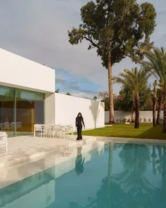 A modern outdoor living space with a pool surrounded by a white wall, a patio with a dining table and chairs, and potted plants, with a person standing near the pool, lit by natural sunlight