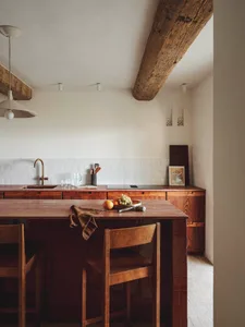 Modern kitchen with a wooden countertop, dining table, and chairs, featuring a sink, refrigerator, and bowl, with a wooden floor and tiled wall, lit by natural sunlight