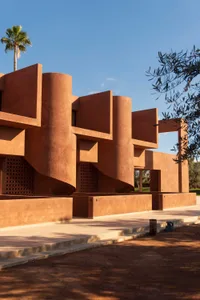 A serene outdoor space with a wooden bench under a tree, surrounded by a modern building with red and brown colors and a large window