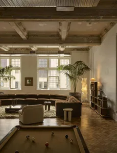 A modern living room with a pool table, large couch, chair, dining table, and potted plants, filled with natural light from multiple windows