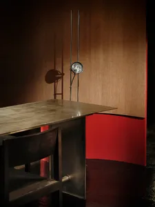 A modern dining room with a metal table and wooden chairs, against a red wall, with dim lighting creating a cozy atmosphere