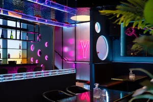 A modern restaurant interior with a black and pink color scheme, well-lit with a combination of natural and artificial light, featuring a sleek bar area with a sink and bottles