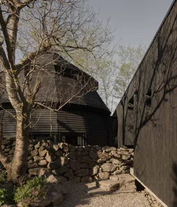 A black house with a stone wall and a round structure, surrounded by natural light and shadows