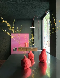 A modern living area with a dining table, red vases, and flowers, surrounded by chairs and a potted plant, with a warm and inviting ambiance