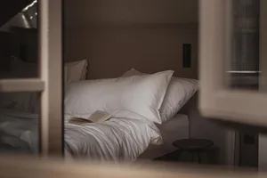 Cozy bedroom with a bed, chair, and book, lit by natural light from the window, with wood and fabric visible
