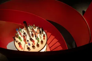 A spiral staircase with a red wall and a large number of bottles displayed in various positions, creating a lively and bustling environment