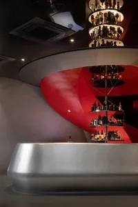 A stylish bar with a red spiral staircase, metal staircase, and a circular display, surrounded by bottles and wine glasses on shelves