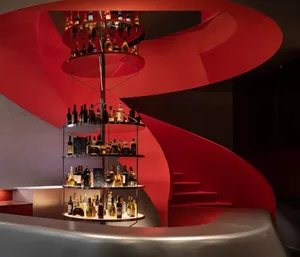 A dimly lit bar with a red metal spiral staircase and a shelf filled with liquor bottles, creating a sophisticated and inviting atmosphere