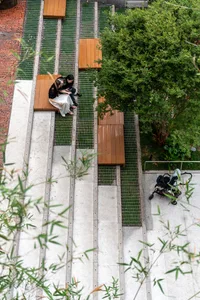 A woman sits on a wooden bench near a tree in a park, with another person standing nearby, creating a peaceful outdoor scene