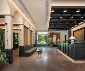 A modern hotel lobby with a large couch, bench, and dining table, surrounded by potted plants and illuminated by natural light