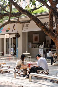 A casual outdoor dining area with people sitting at tables and chairs under a tree, surrounded by a modern restaurant with wood and metal materials, lit by natural sunlight