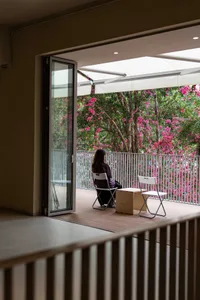 A woman sits on a white chair on a wooden deck surrounded by a garden with pink flowers, enjoying the peaceful atmosphere