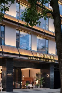 飯店入口處有大型窗戶、招牌與盆栽，背靠現代建築的混凝土與玻璃立面，自然陽光沐浴其中