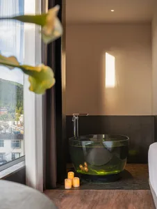 A dimly lit bathroom with a large green-tinted bathtub, sink, and toilet, surrounded by potted plants and a city view through the window