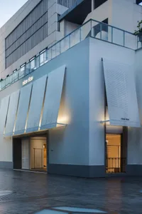 Photograph of a modern building with a white concrete facade, a large storefront window, and vertical blinds, illuminated by a light source