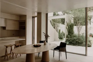 A modern living room with a dining table, chairs, and a potted plant, surrounded by a large window allowing natural light to enter the space