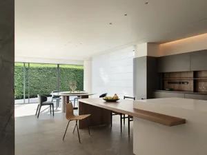 Modern kitchen and dining space with a large island featuring a marble countertop, a sink, oven, and refrigerator, surrounded by chairs and a dining table, with a potted plant and vase adding greenery and decoration