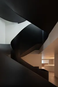 A dark and moody photograph of a spiral staircase with wooden treads and a metal railing, set against a white wall