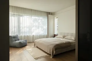 A modern bedroom with a bed, chair, and window, featuring a predominantly white color scheme and natural light from the window