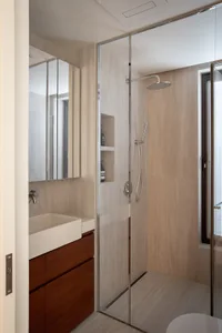 Modern bathroom with a glass shower, wooden cabinet, and white sink, surrounded by tiled walls and natural light