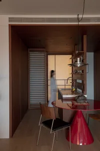 A modern kitchen with a wooden floor, a red dining table, and a variety of furniture and items, lit by natural sunlight