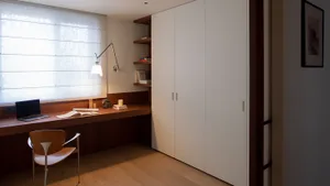 A cozy bedroom with a wooden floor, a white cabinet, a bed, a chair, a desk, and a book, creating a warm and inviting atmosphere