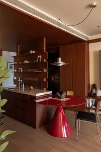 A modern kitchen with wooden cabinets, a red dining table, and a potted plant, surrounded by chairs and natural light from the windows