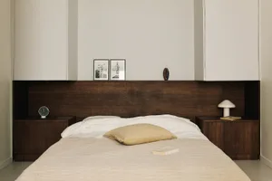 A calm and inviting bedroom with a white comforter, a tan pillow, a nightstand with a lamp, and a dresser in the corner, set against a clean and well-organized white-walled space