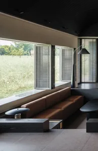 A modern living room with a brown couch, a black chair, and a dining table, well lit with a large window