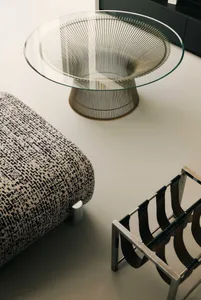 A well-lit living room with a circular glass table, a metal-framed chair, a couch, and a vase, featuring a modern style and sleek metal and glass materials