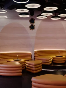 A modern lounge with a circular layout, featuring a central table surrounded by semi-circular couches, illuminated by natural and artificial light, creating a warm and inviting atmosphere