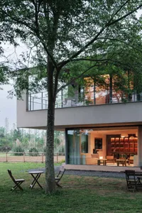 A modern house with a glass wall and large window, featuring a patio with a dining table and chairs, and a dining area with a couch and chair, illuminated by natural light