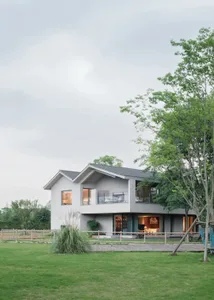 A modern house with a large white exterior, surrounded by a lush green field, featuring a deck and patio area with a bench and dining table, and a well-lit interior with comfortable furniture