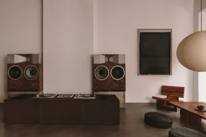 A cozy living space with a wooden floor, a white wall, a wooden speaker, a bench, and a dining table with a chair, creating a warm and inviting atmosphere