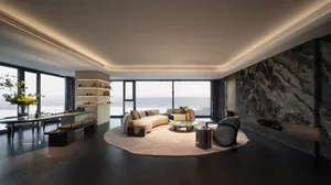 海濱客廳呈現現代美學，擁有大型窗戶飽覽海景，搭配圓形地毯、沙發、椅子、餐桌與裝飾品，強調開敞感與自然光線