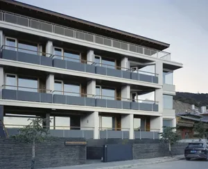 Modern multi-story building with a balcony, black railing, and a backdrop of mountains