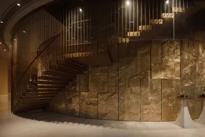 A dimly lit room with a brown stone staircase and wooden railing, set against a large stone wall