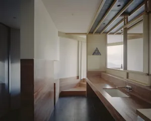 A minimalist bathroom with a wooden bench, sink, and window, featuring a dimly lit, warm, and natural atmosphere