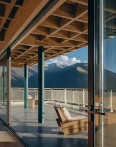 A modern living room with a wooden floor, a wooden chair, and a couch, with a large glass door providing a view of the mountains outside, and natural light streaming in