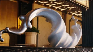A modern metal sculpture is placed in a museum or art gallery setting with a potted plant nearby, illuminated by natural sunlight